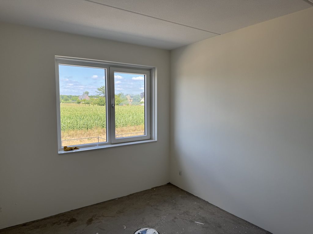 Lege nieuwbouwkamer met strakke witte wanden, een kale vloer en uitzicht op een maisveld