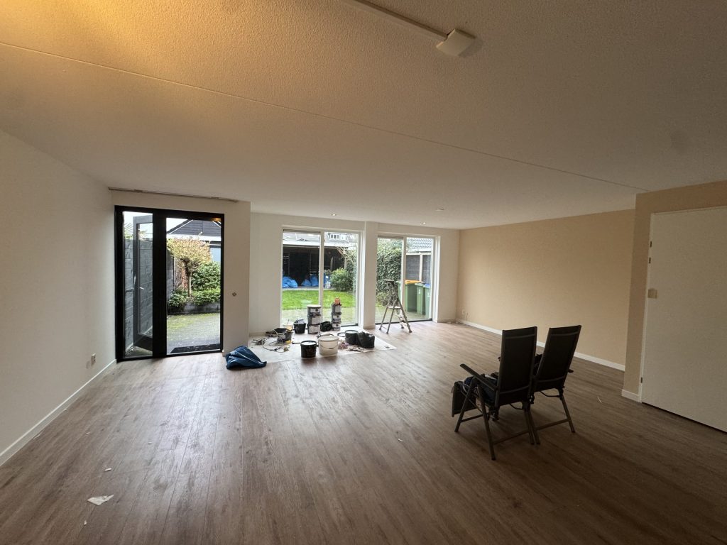 Woonkamer van een bestaande woning met lichte wanden, een houten vloer en verfemmers bij de tuindeuren